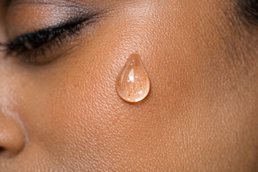 Close-up of a clear drop of face serum on medium-brown Indian skin, just before absorption.