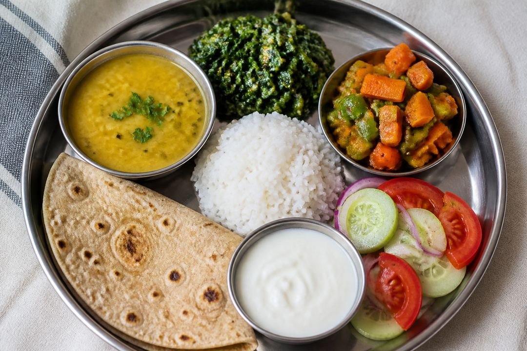 Balanced Indian thali on a stainless steel plate with roti, a small portion of rice, dal, leafy green sabzi, a colourful vegetable sabzi, salad and a bowl of curd.